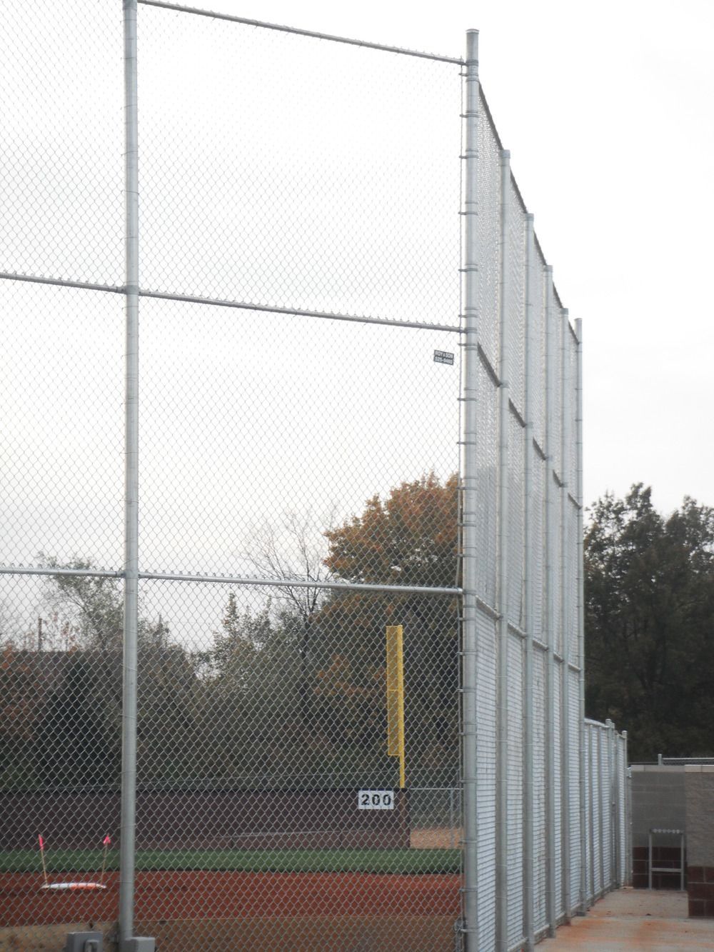 Chain-link fence around a baseball field with a yellow foul pole and trees in the background.