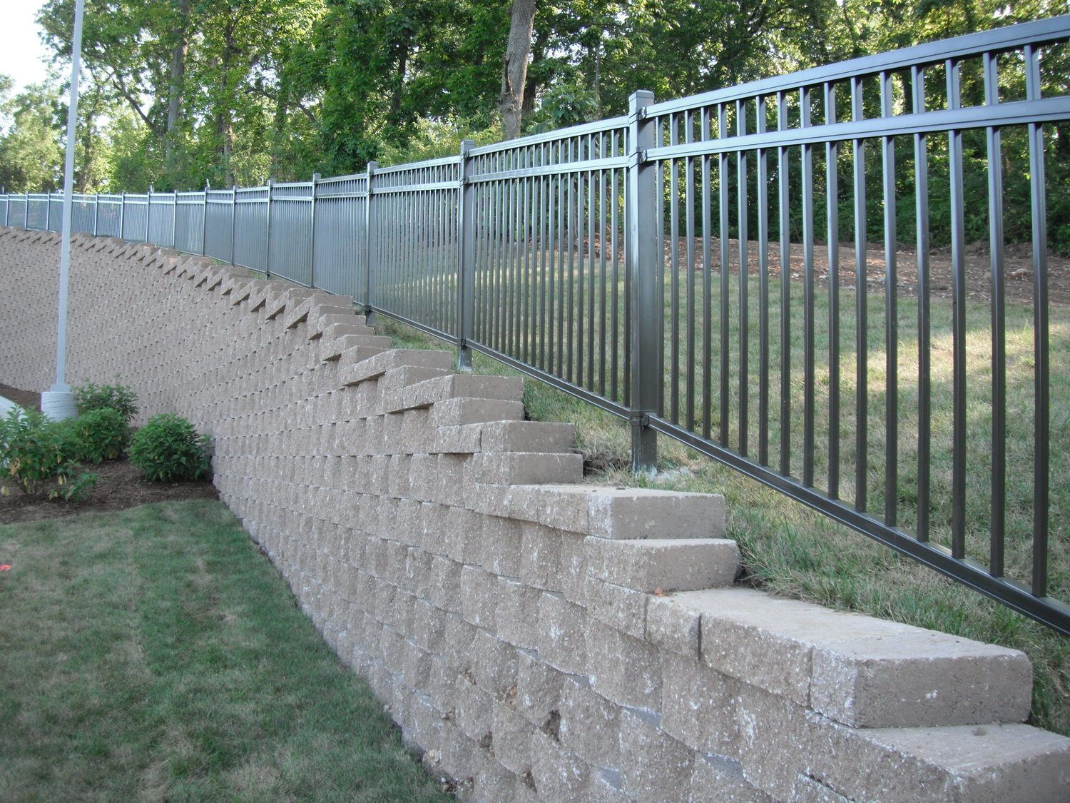 A metal fence atop a retaining wall built of gray blocks, on grassy lawn.
