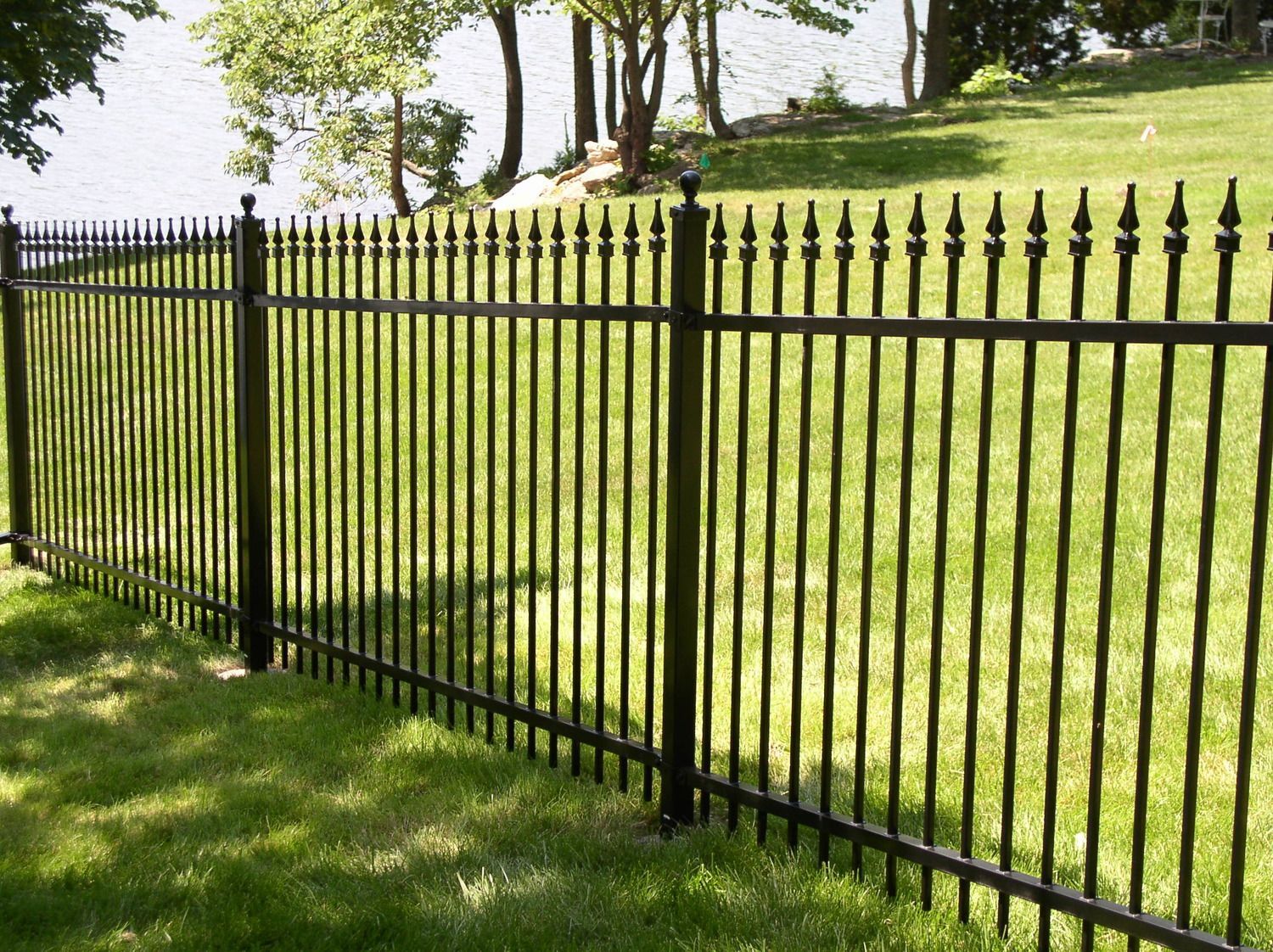 Black metal fence bordering a grassy lawn, with water and trees in the background.