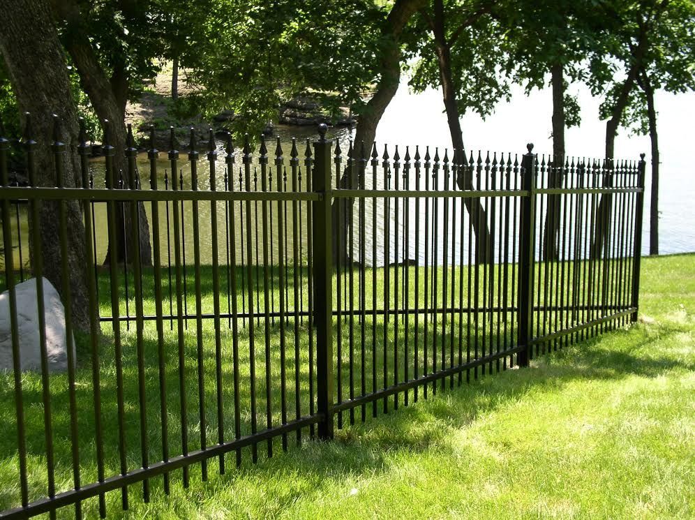 Black metal fence bordering a grassy lawn, trees, and a body of water.