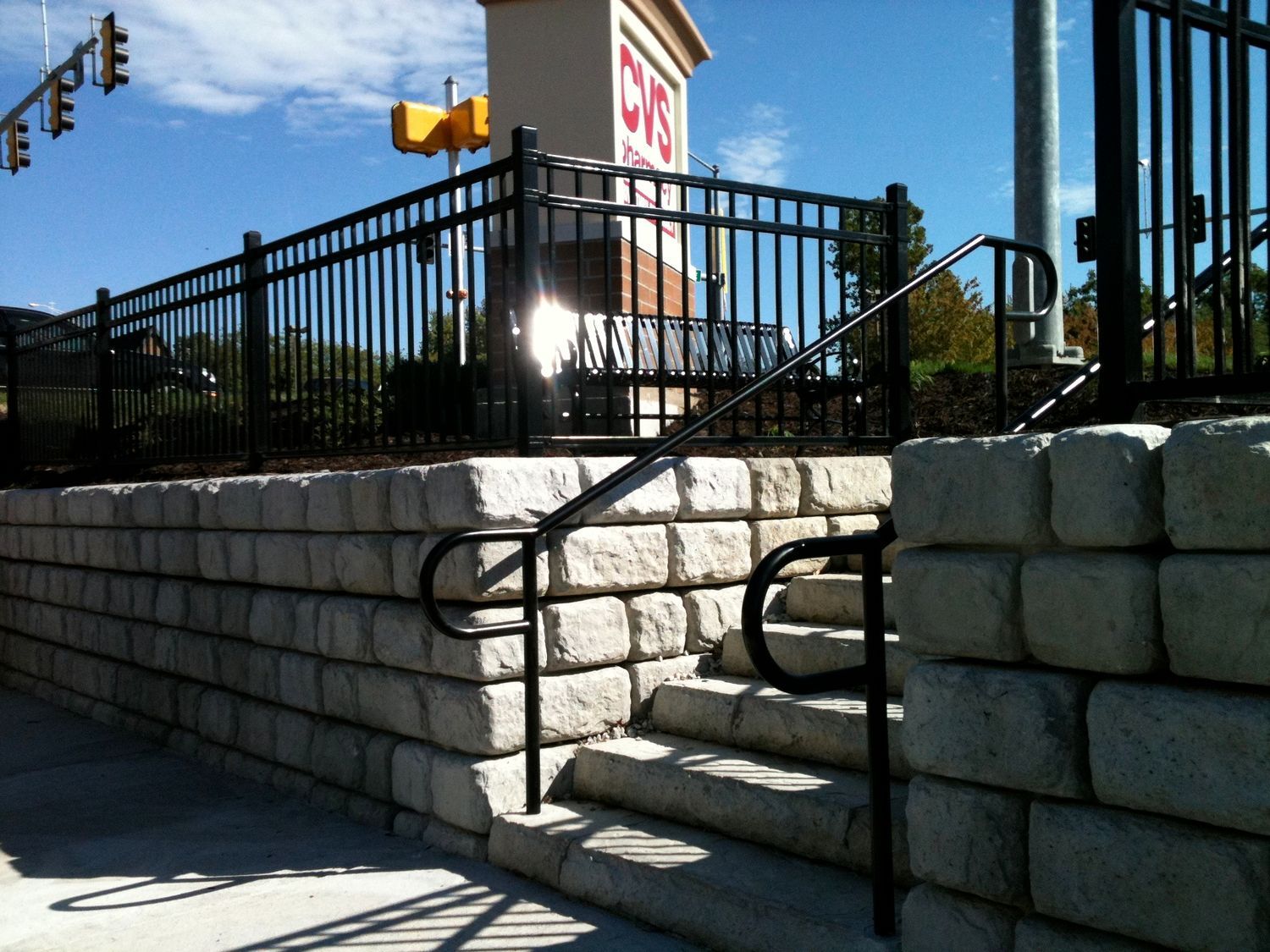 A stone retaining wall with steps, a black handrail, and a black fence. In the background is a restaurant under a blue sky.
