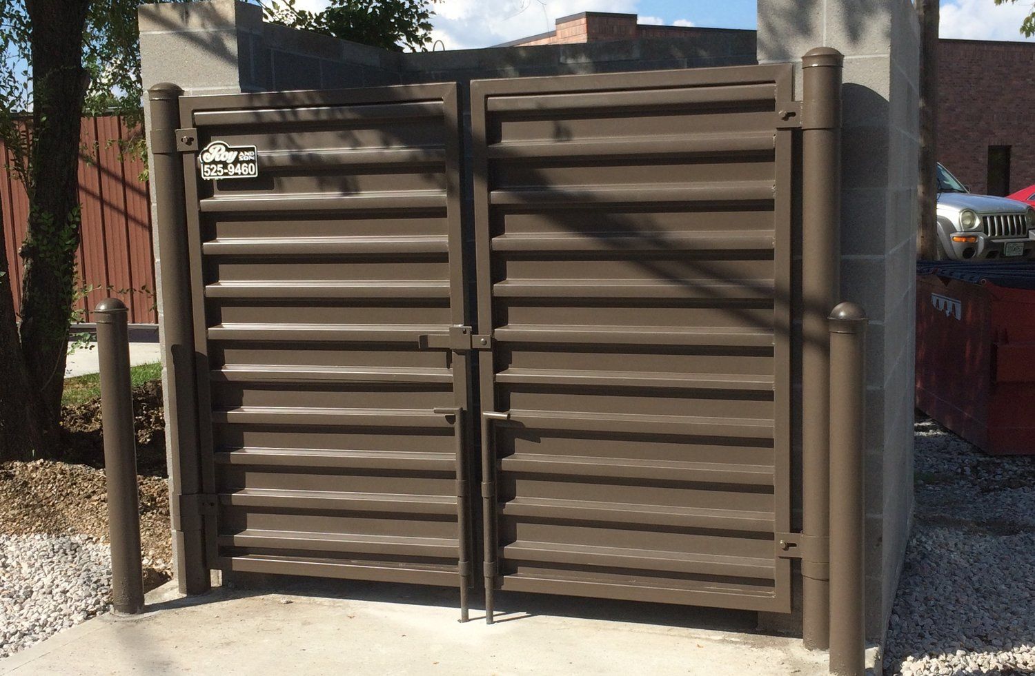 Brown metal dumpster enclosure with closed gate on a concrete pad.