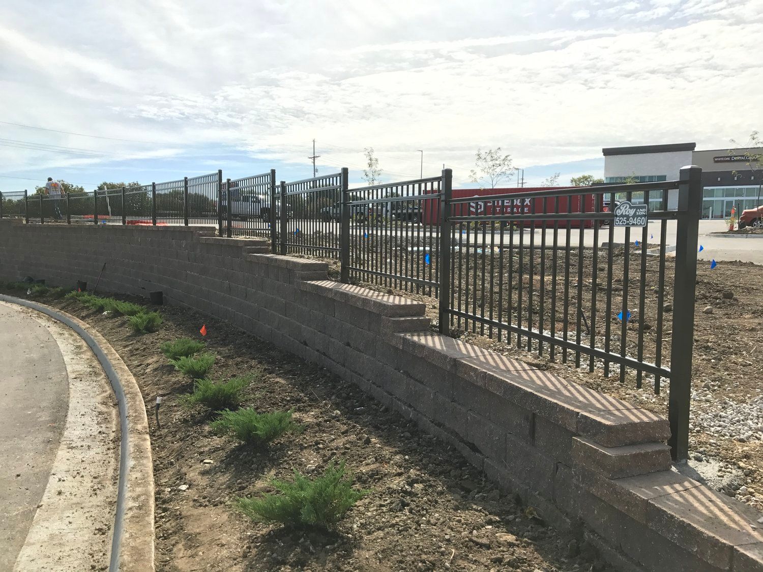 Black metal fence on a brick retaining wall, with landscaping and construction in the background.