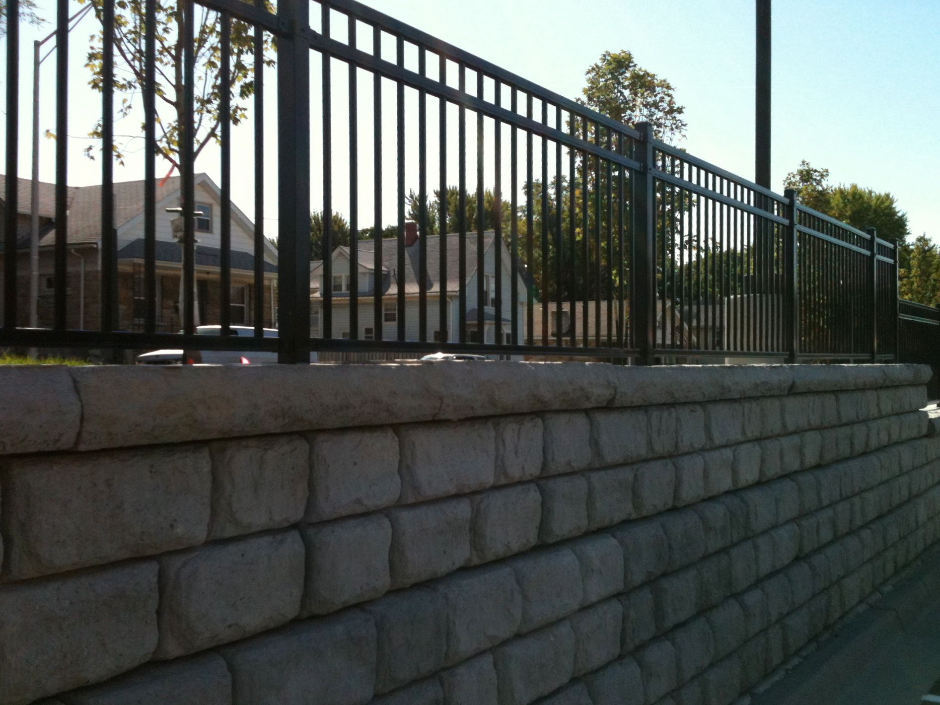 Black metal fence atop a stone retaining wall, with houses and trees in the background.