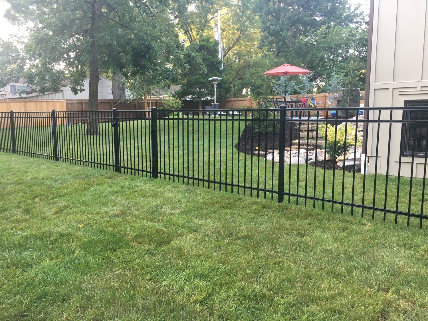 Black metal fence encloses a green lawn, trees, and a house exterior.