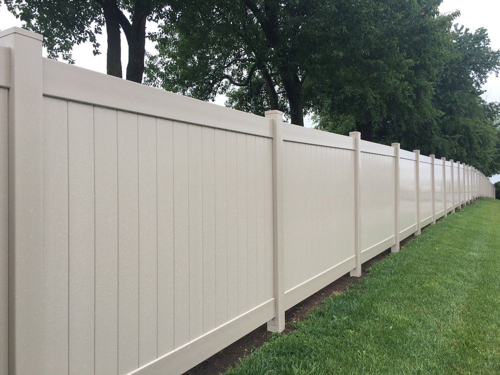 Tan vinyl privacy fence along green grass, trees in background.