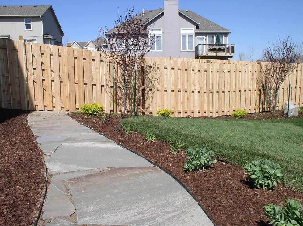 Stone path leads through a backyard with a wooden fence, green grass, and small bushes.