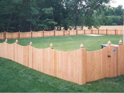 Wooden fence with scalloped top and finials encloses a grassy yard, trees in background.