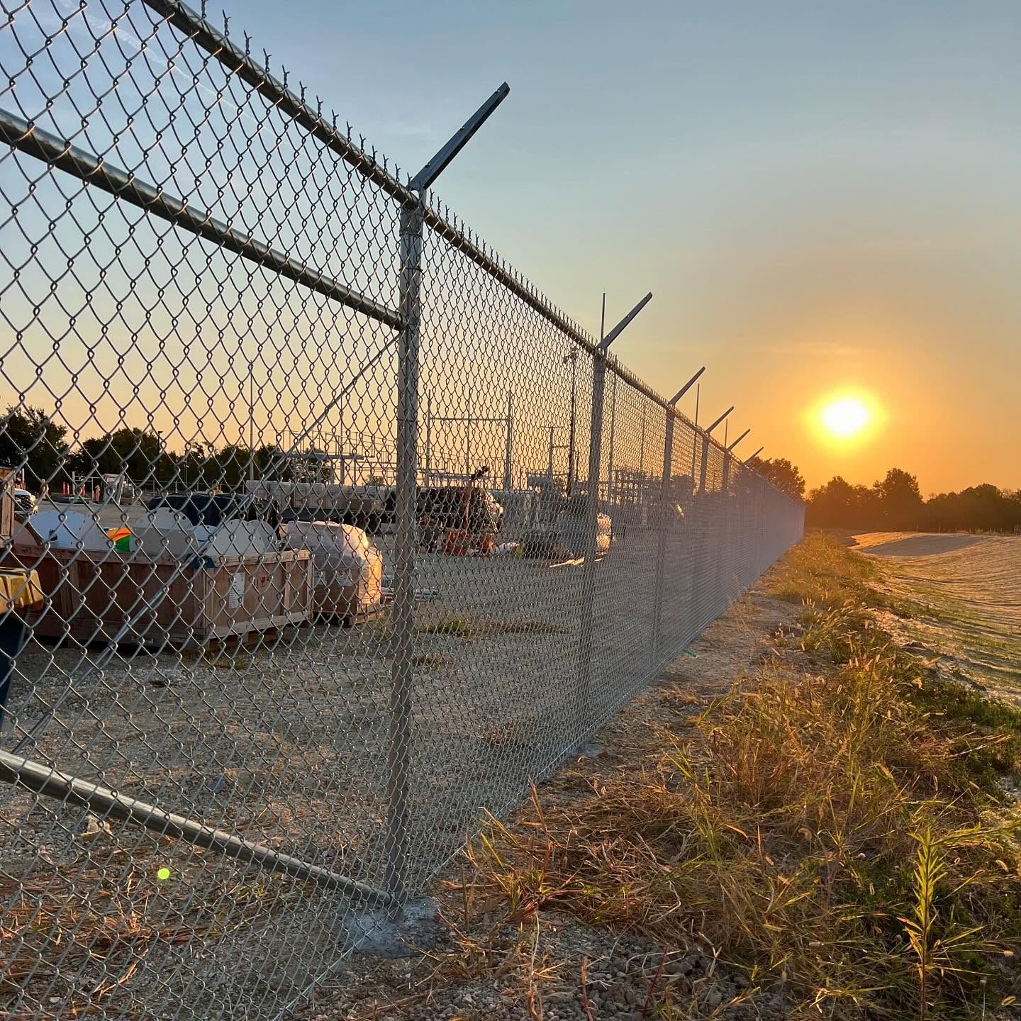 Chain-link fence with barbed wire topping, silhouetted against a sunset.