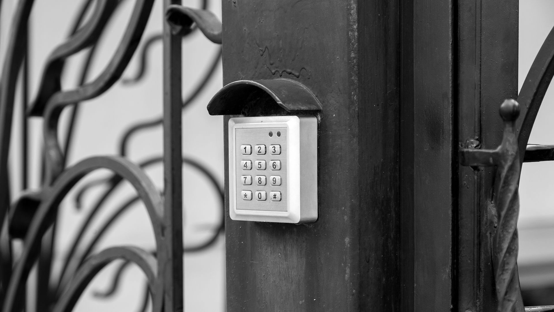Black and white shot of a keypad mounted on a black metal gate post, sheltered by a small hood.