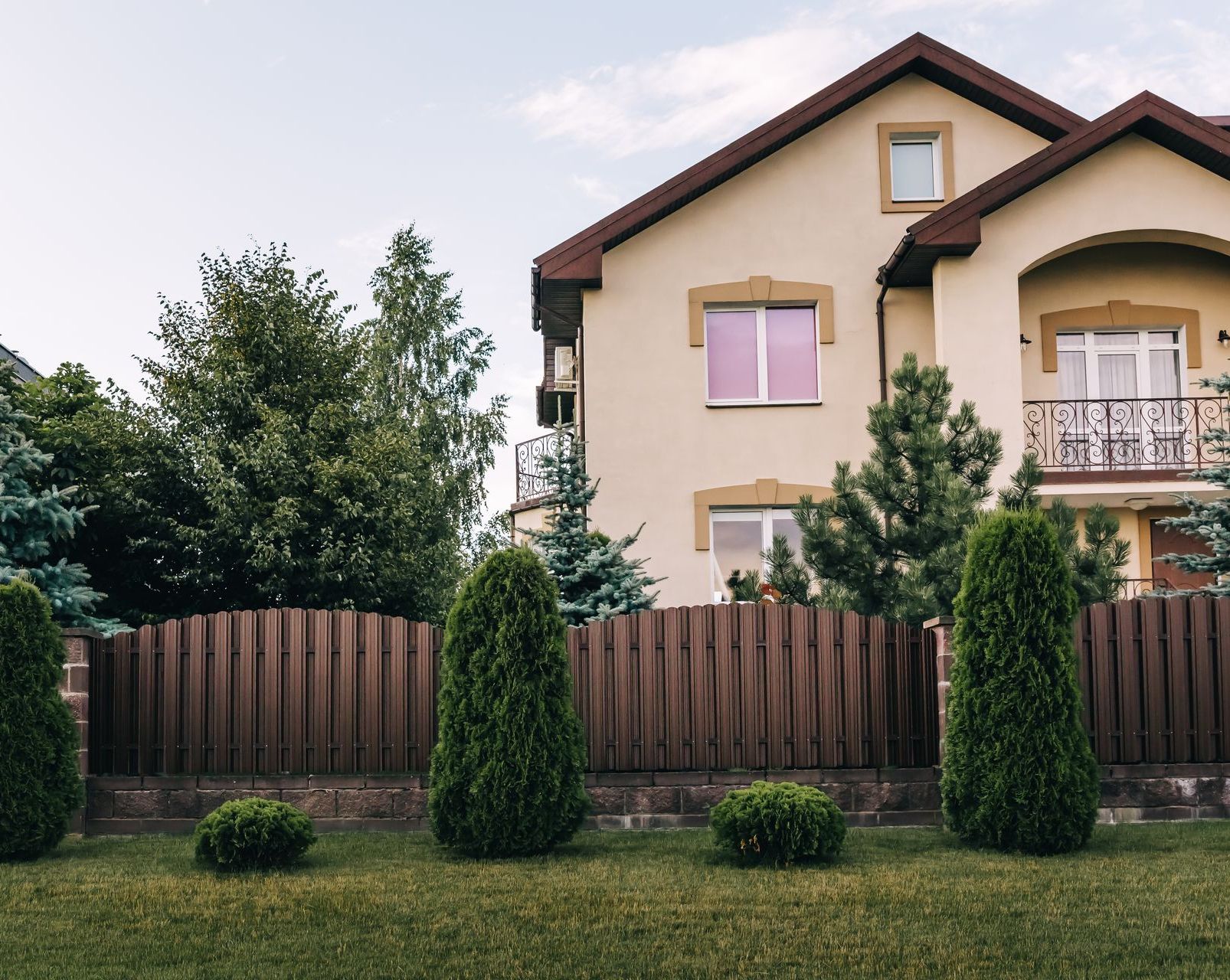 Tan house behind a brown fence and trimmed bushes. Lush green lawn and trees under a cloudy sky.