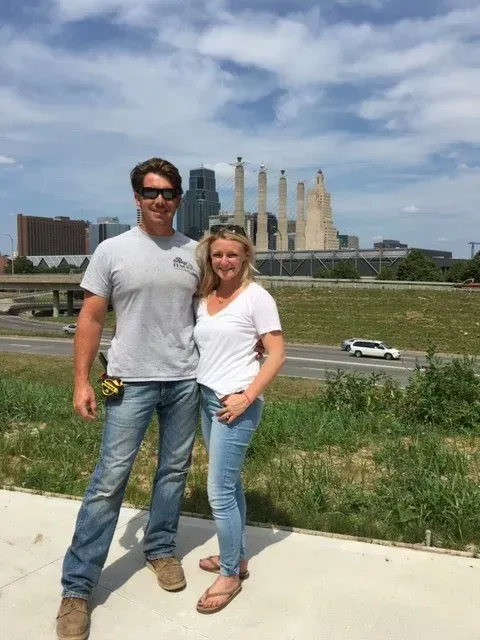 Man and woman pose in front of a city skyline and memorial sculpture under a blue sky.