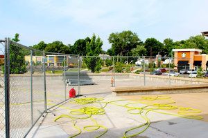 Empty, fenced-off pool with yellow hoses, red fire extinguisher, and buildings/trees in background.