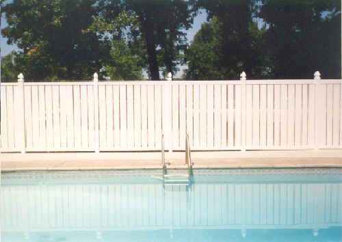 White fence surrounds a pool with a ladder; trees in the background, blue water.