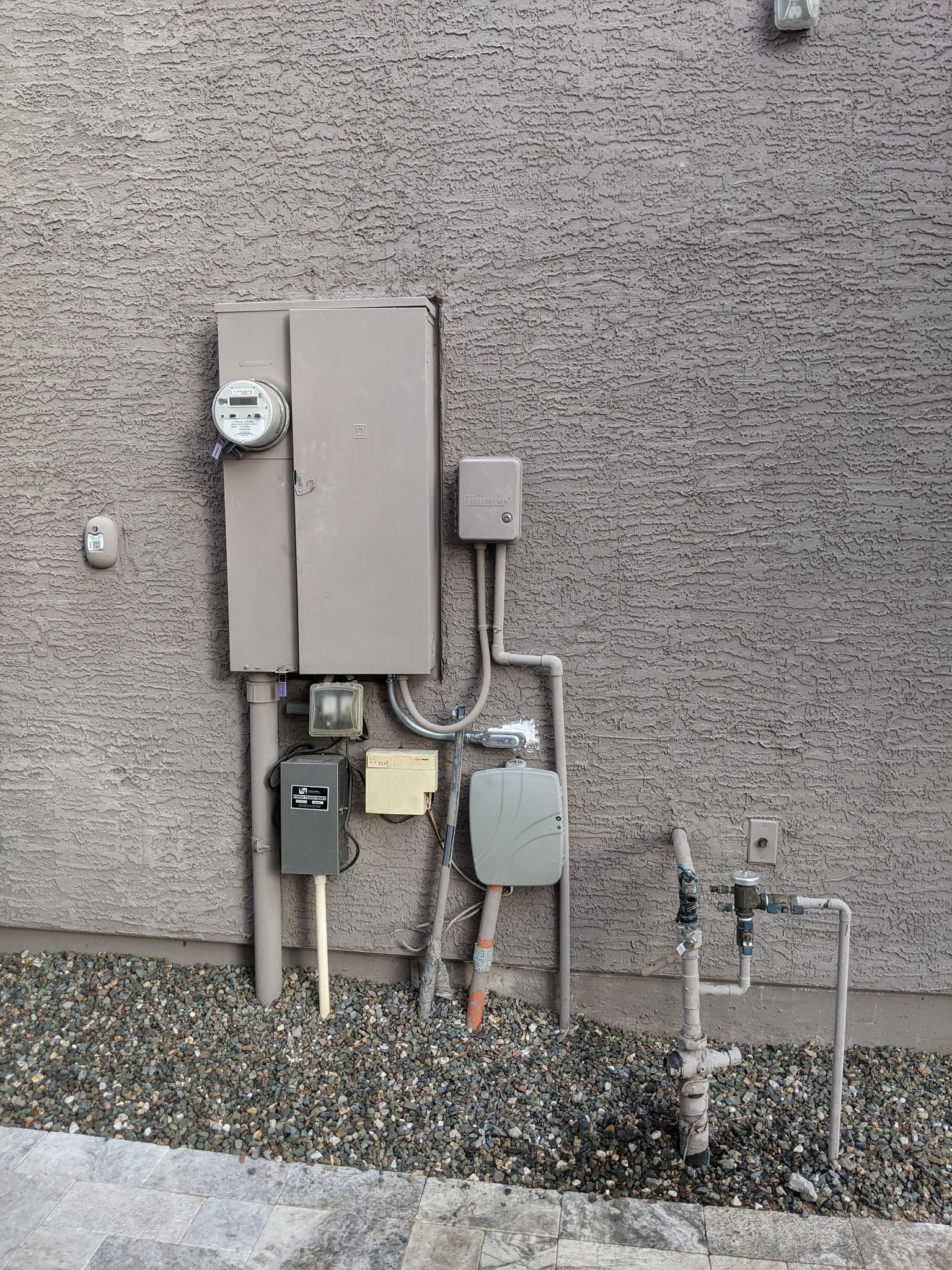 An electrical meter, breaker box, and plumbing pipes mounted on the exterior of a stucco wall above a gravel ground.