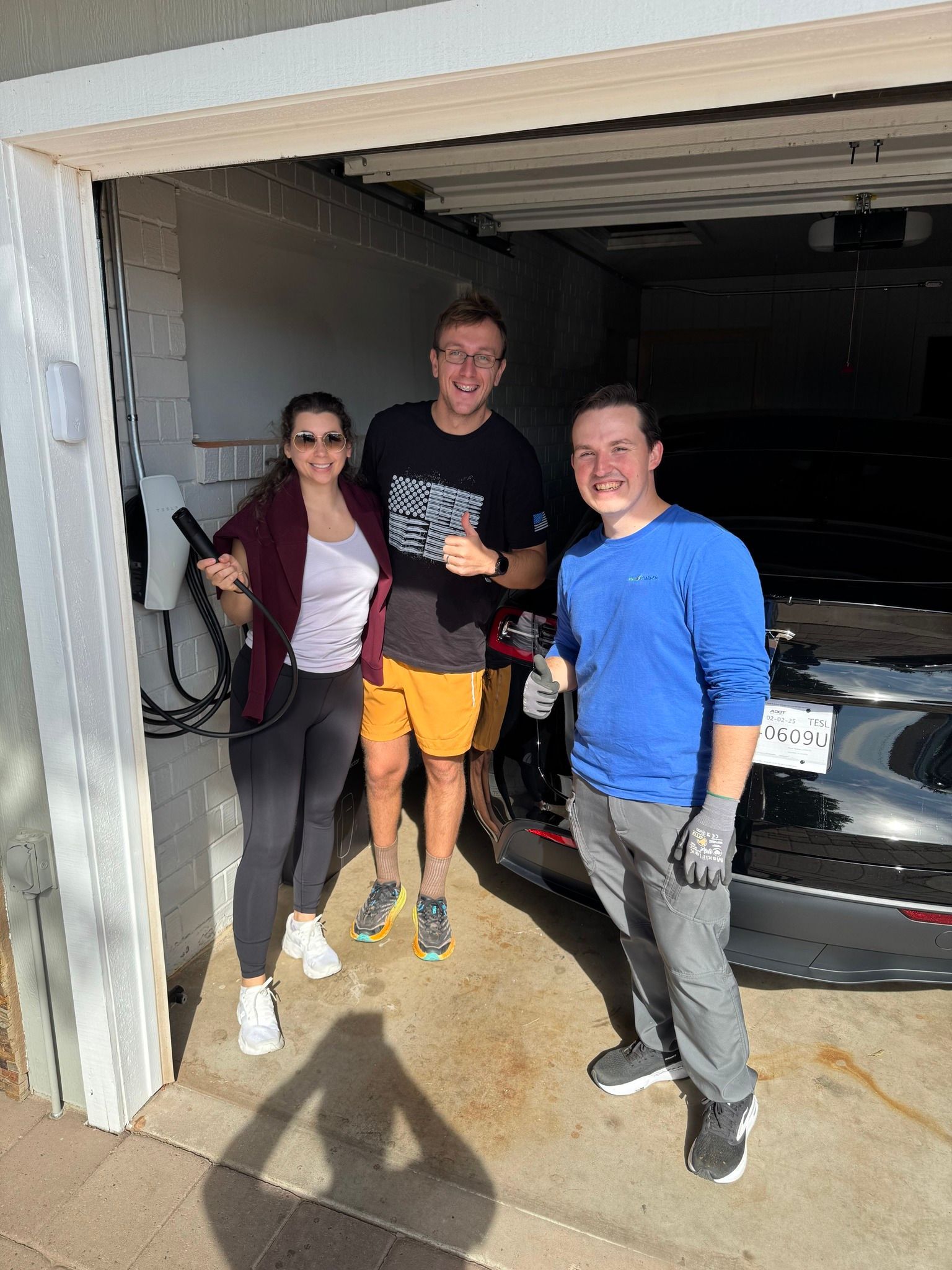 Three people stand in a garage next to an electric vehicle charging station, smiling near a black car.
