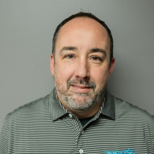 Man with short brown hair and beard smiles, wearing a striped polo shirt against a grey background.