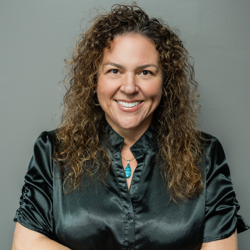 Woman with curly brown hair smiles wearing a black satin shirt with a turquoise pendant.