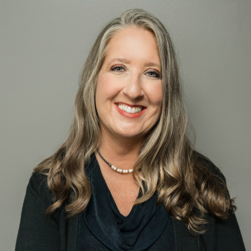 Woman with long, wavy gray hair smiles, wearing a navy blouse and pearl necklace, against a gray background.