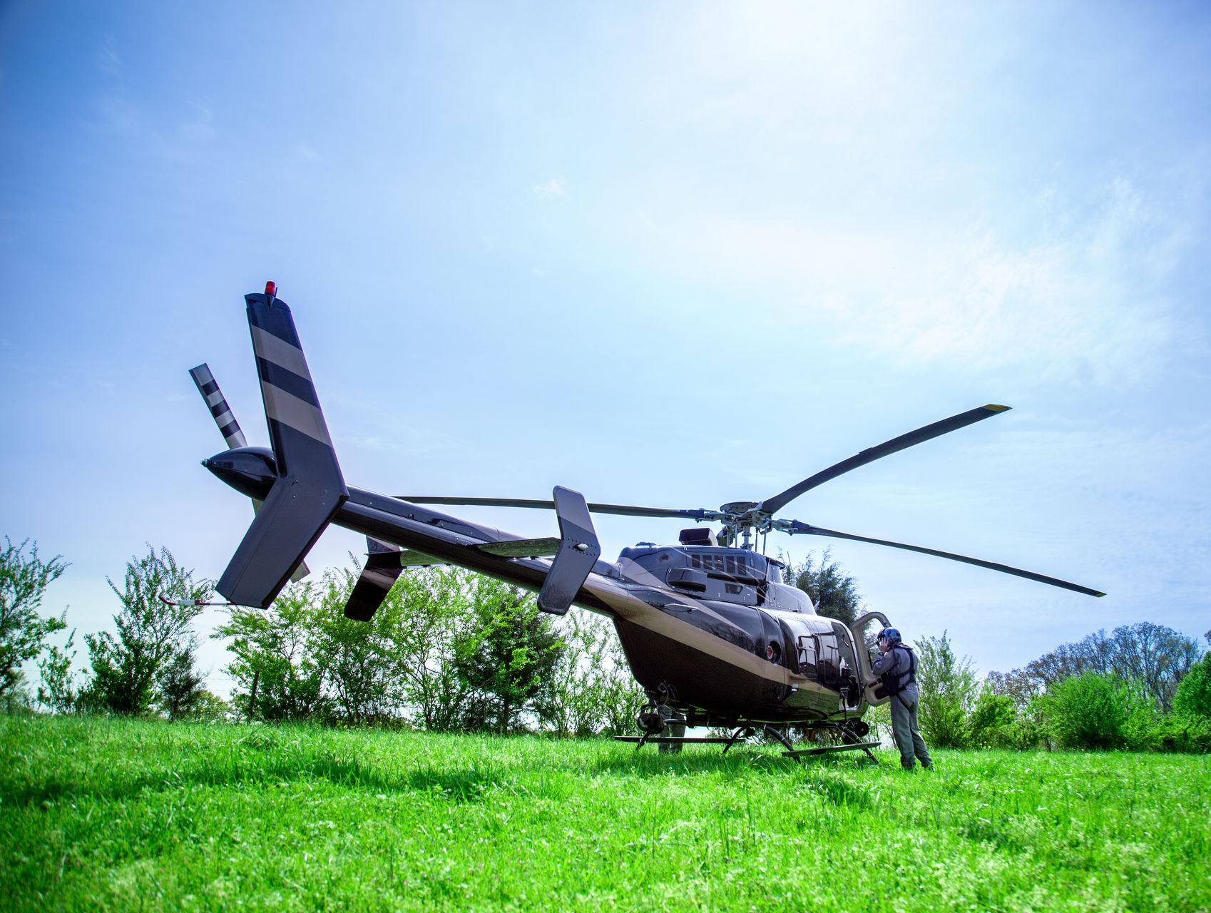 Black helicopter parked on a grassy field on a sunny day. Person stands by the side.