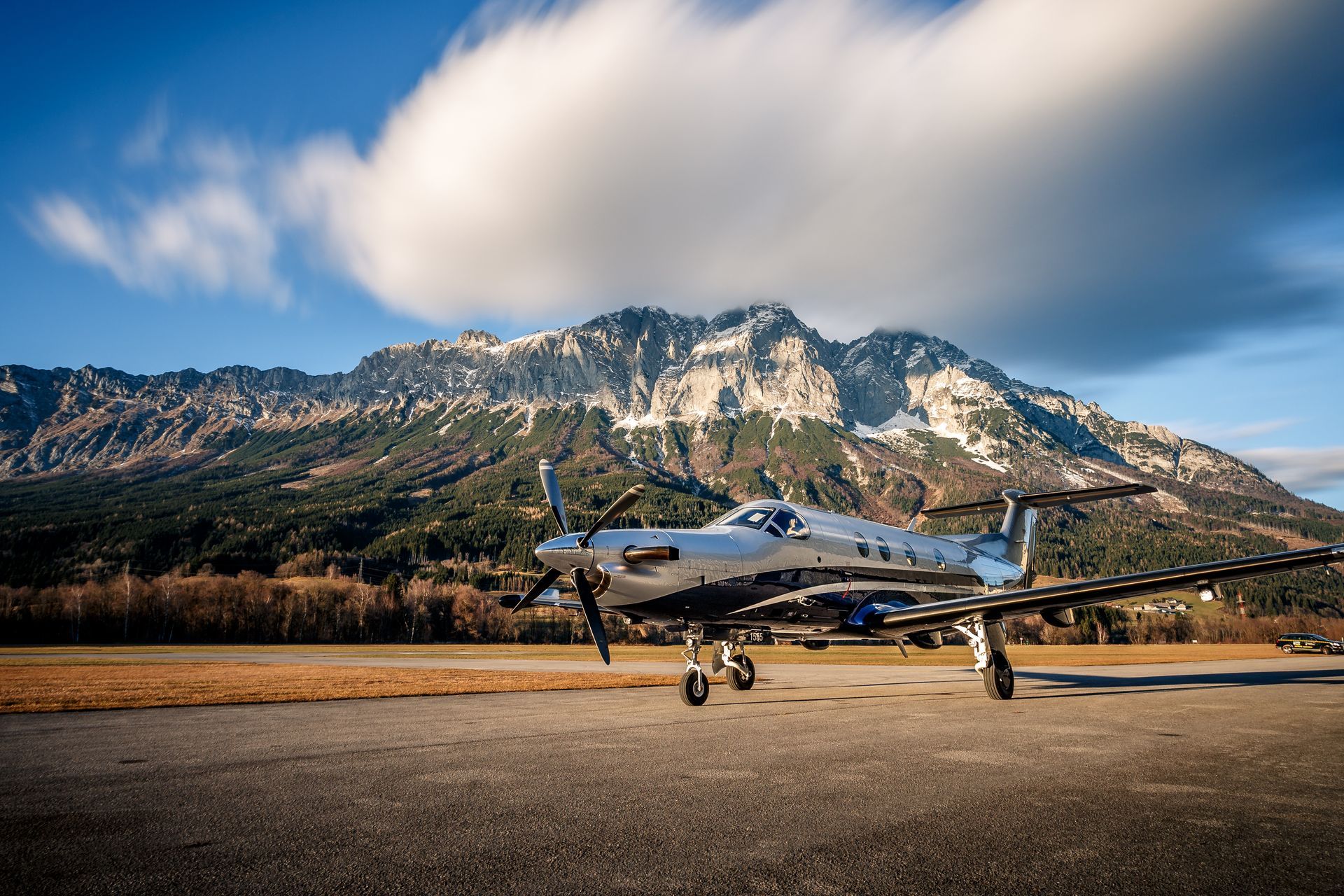 Silver propeller airplane parked on runway, mountains and clouds in the background.