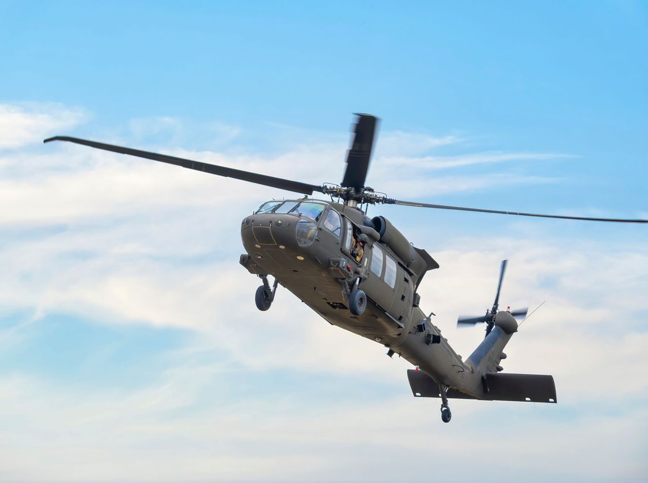 Black military helicopter in flight, against a blue sky with clouds.