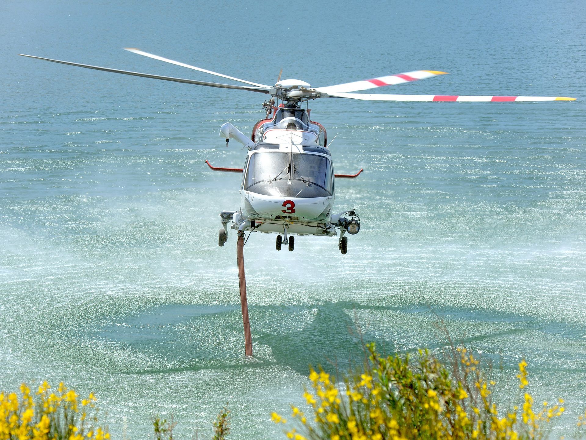 Helicopter with hose collecting water over a body of water, for fire suppression, sunny day.