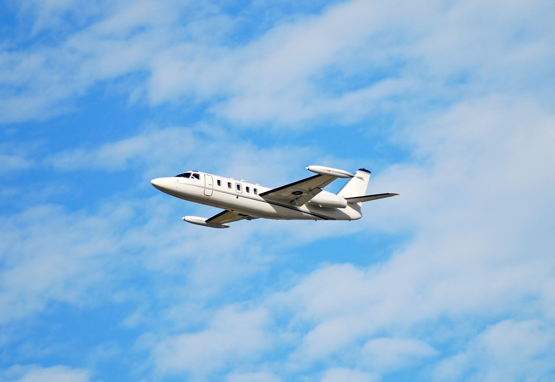 White private jet flying in a blue sky with scattered clouds.