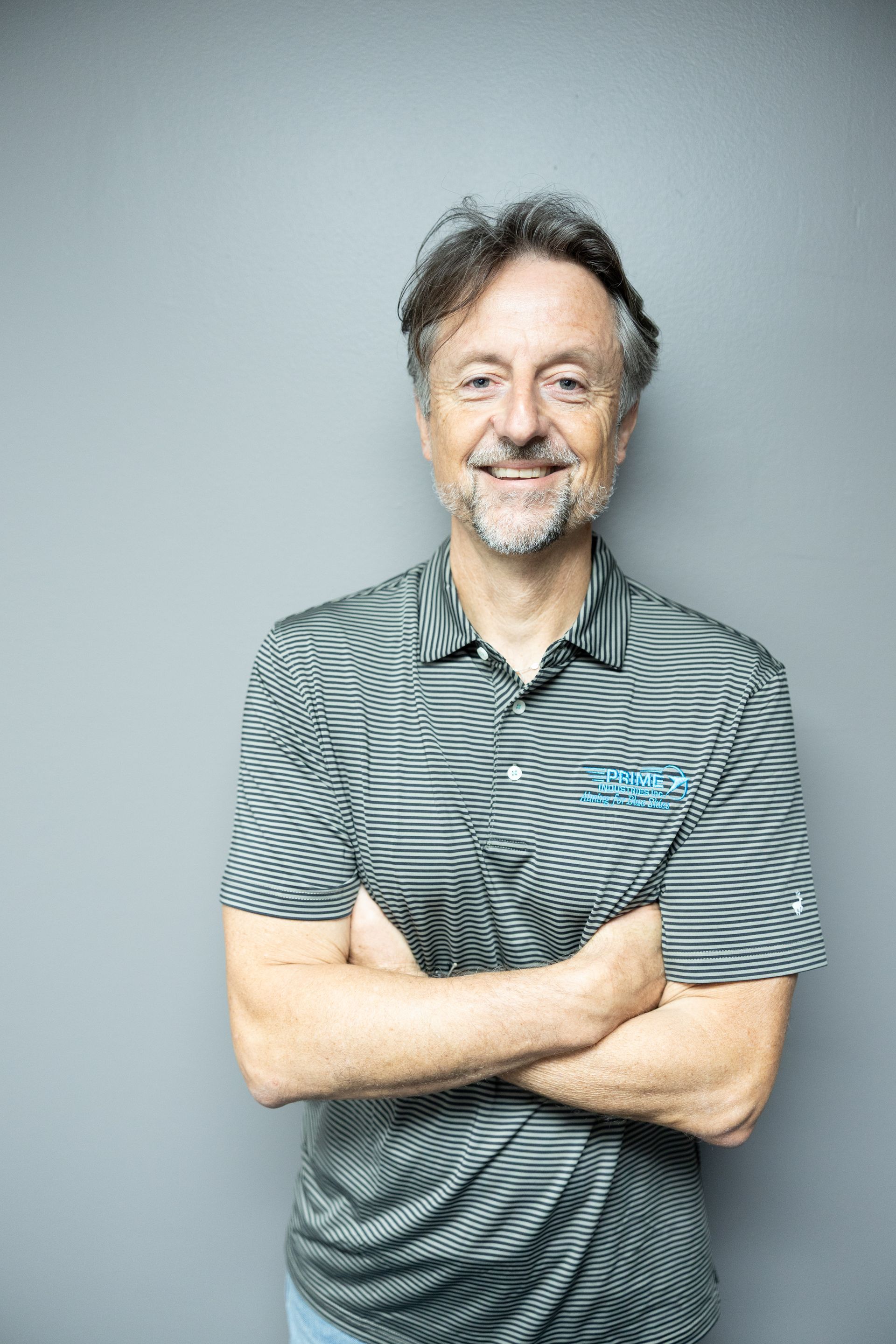 Man with crossed arms, wearing striped shirt, smiling in front of a gray wall.