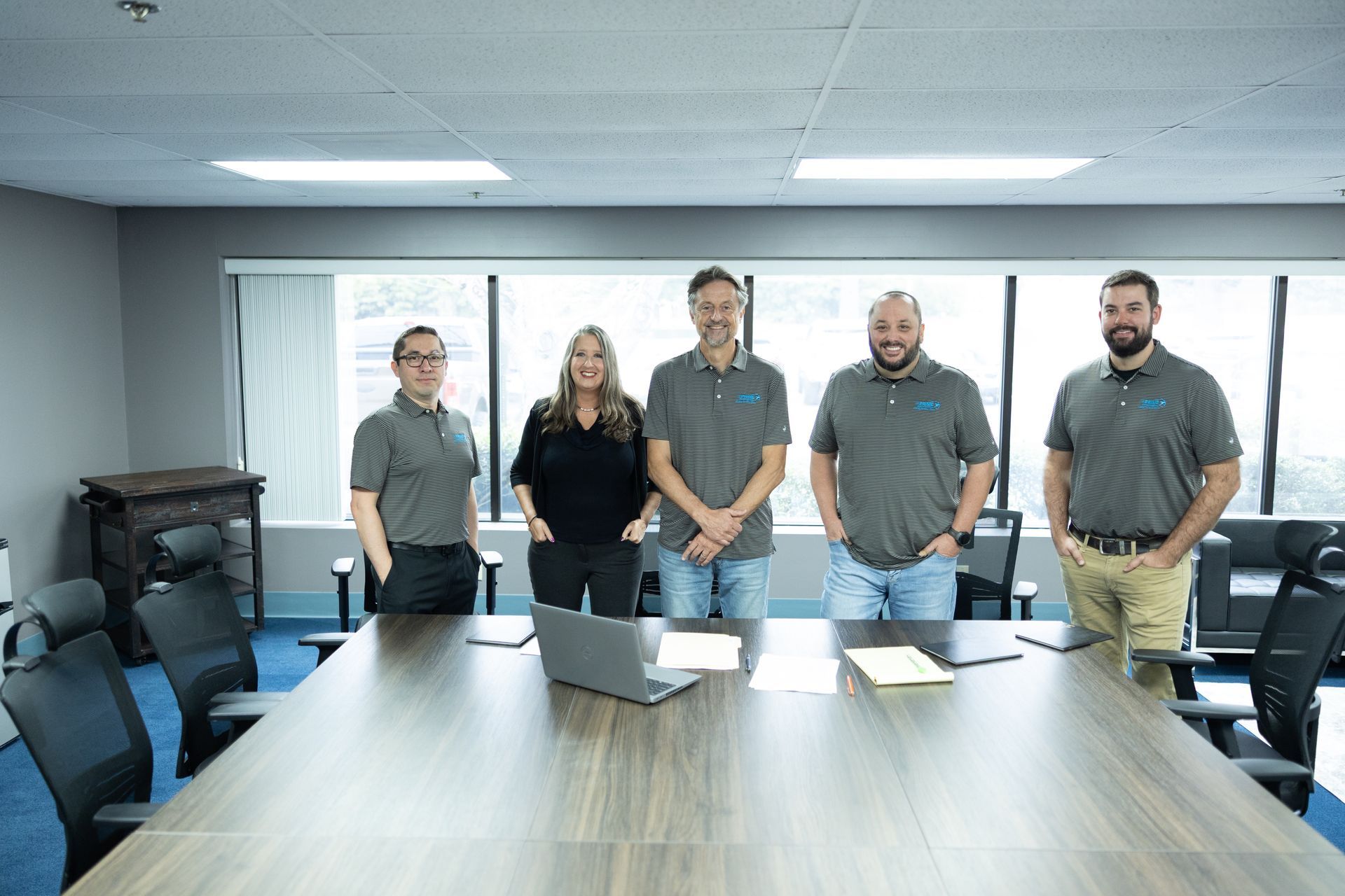 Five people standing behind a conference table. One woman, four men, all wearing polo shirts. Large window in background.