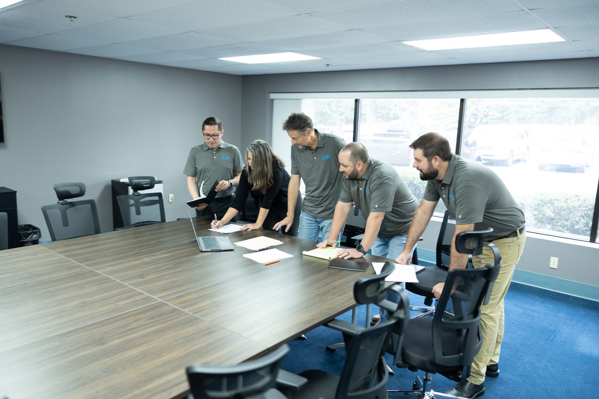 Five people gather around a conference table in an office, reviewing documents and a laptop.