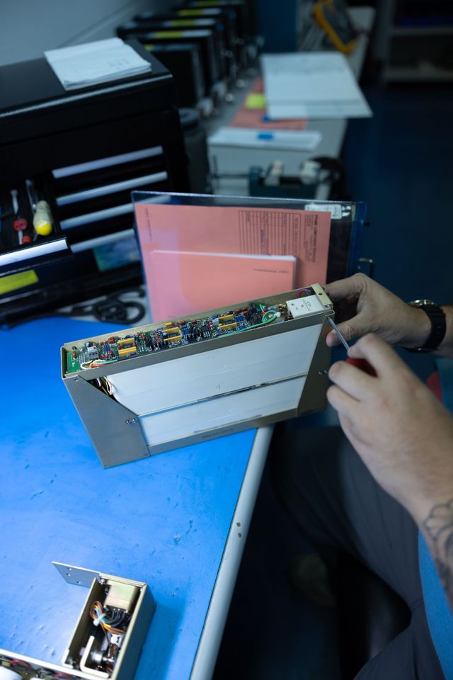 Person using a screwdriver to work on a circuit board inside a metal enclosure on a blue workbench.