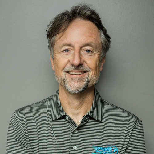 Man with short, graying hair and beard smiles, wearing a striped polo shirt against a gray backdrop.