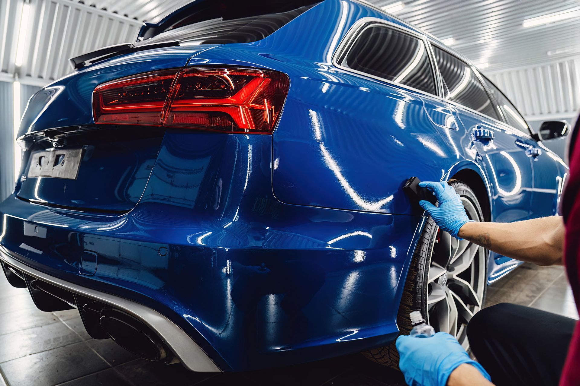 A technician wearing blue gloves polishes the side panel of a vibrant blue station wagon in a bright workshop.