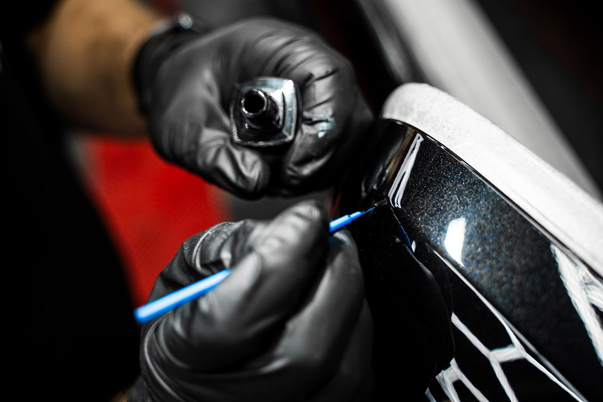 Hands in black gloves use a small blue brush to apply touch-up paint to a scratch on a dark, metallic-finished car surface.