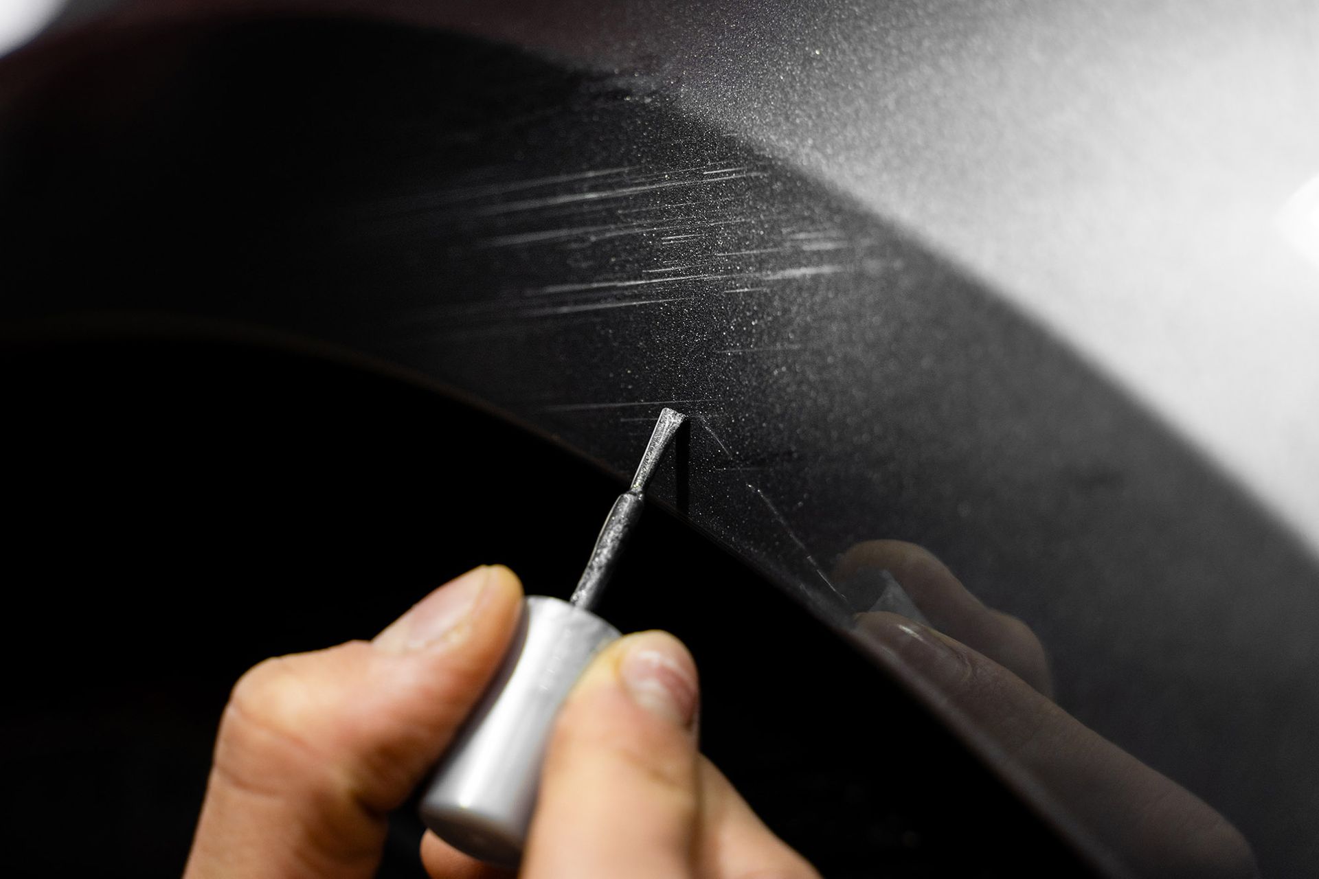 Close-up of a person using a small brush to apply touch-up paint to scratches on a dark car surface.