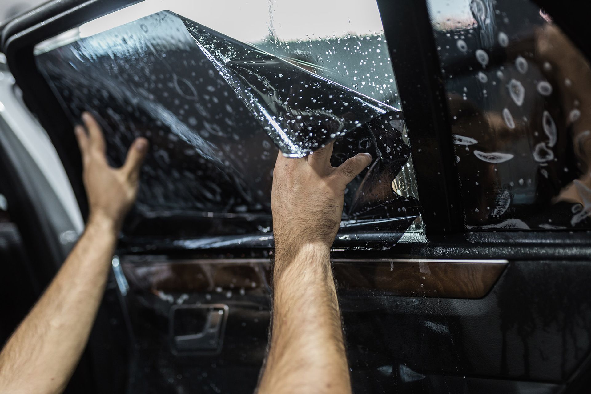 A person carefully applies a dark, wet film to a car's side window.
