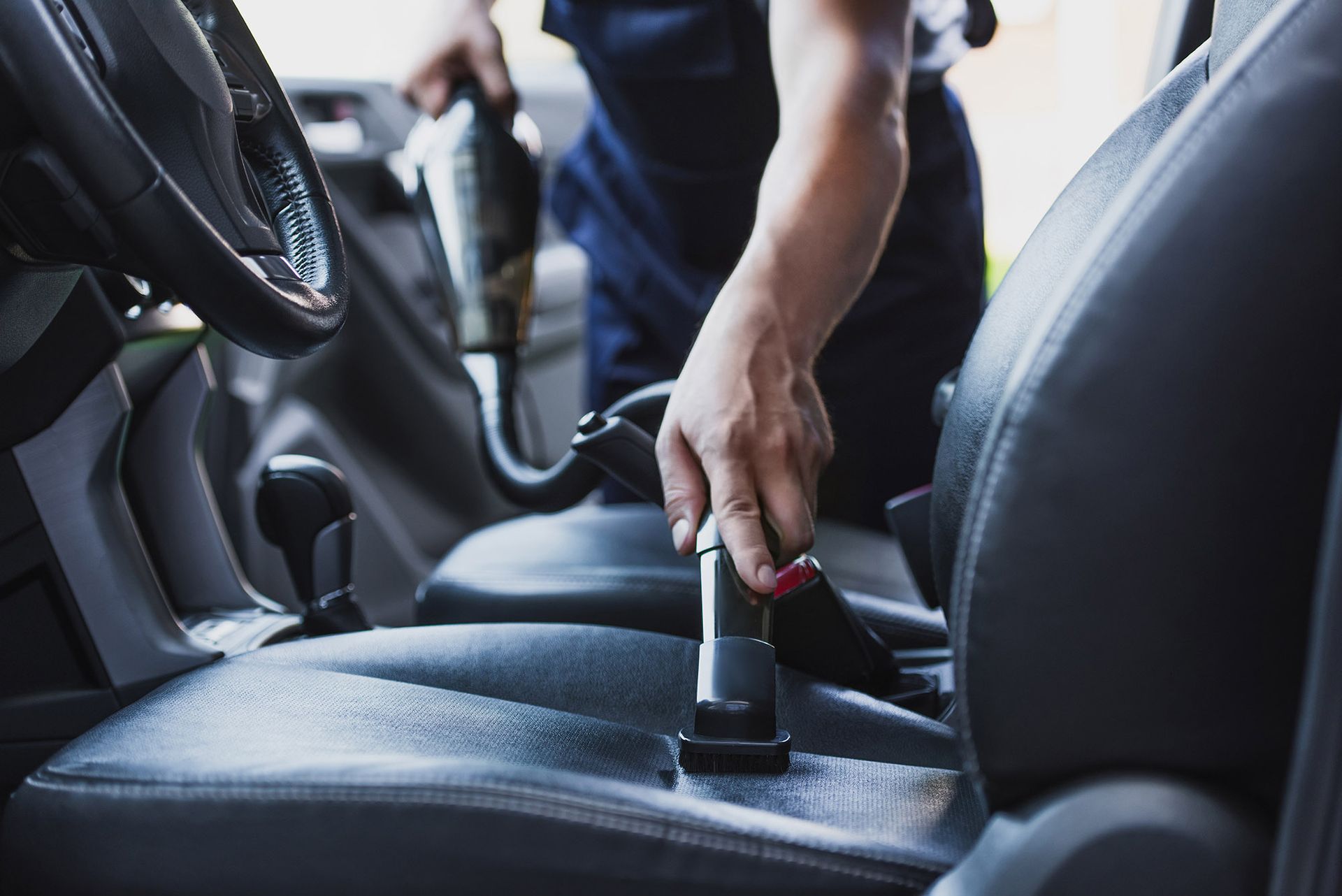 A person vacuums the dark leather seat of a car interior.