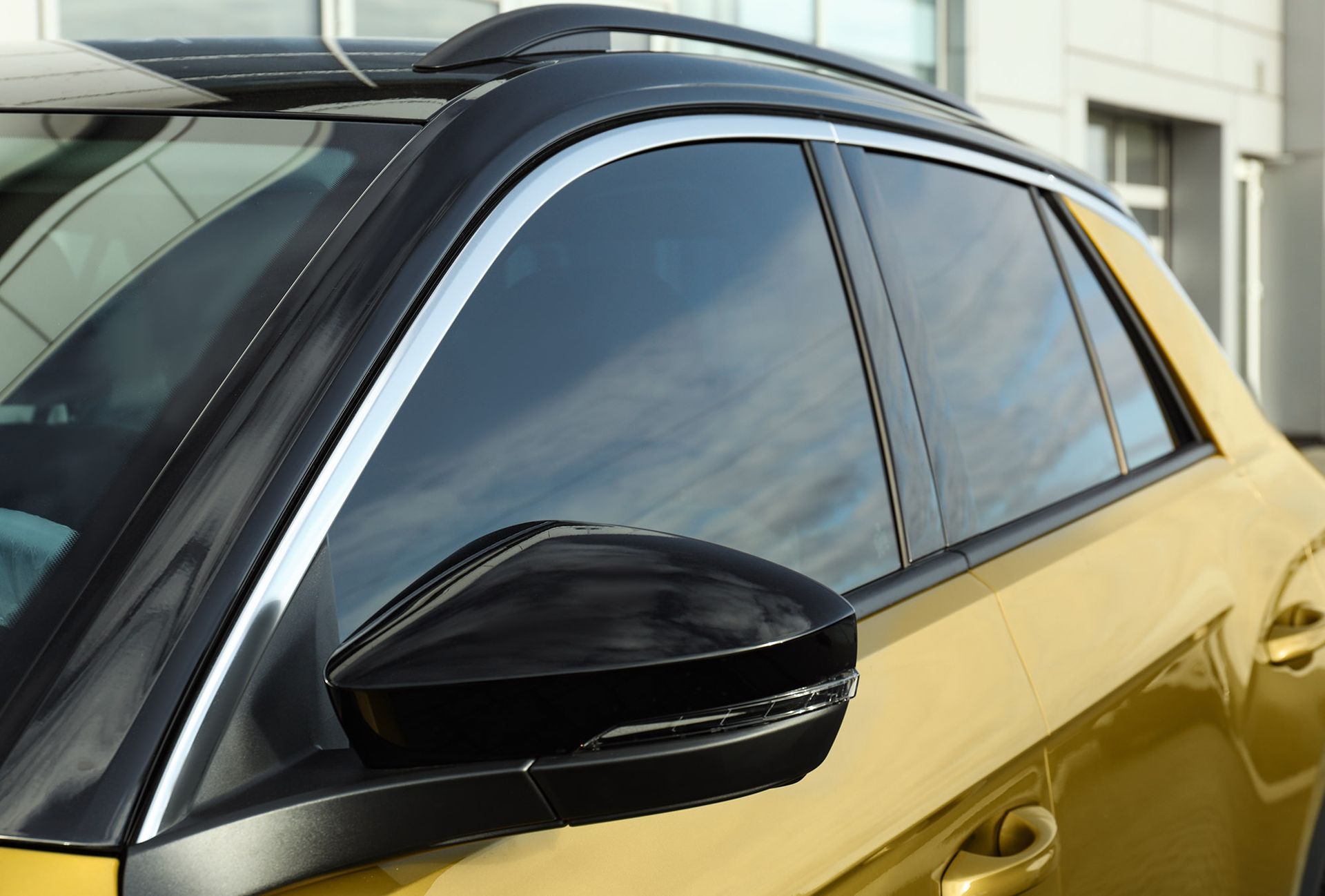 A close-up view of a yellow car's passenger-side door, featuring a black side mirror, window trim, and roof rack.