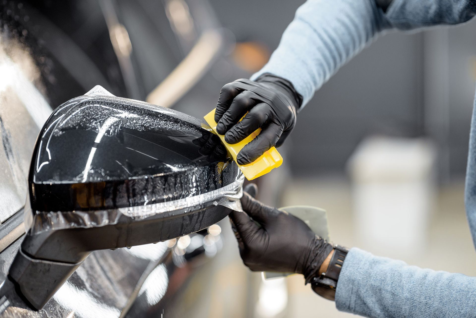 A person wearing black gloves applies a protective film to a shiny black car side mirror using a yellow squeegee.