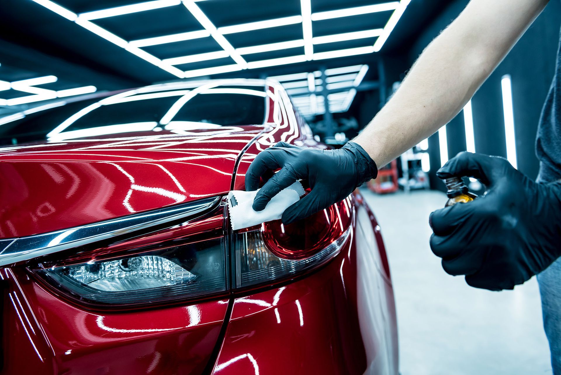 A person wearing black gloves uses a cloth and spray to detail the red tail light of a car in a brightly lit shop.