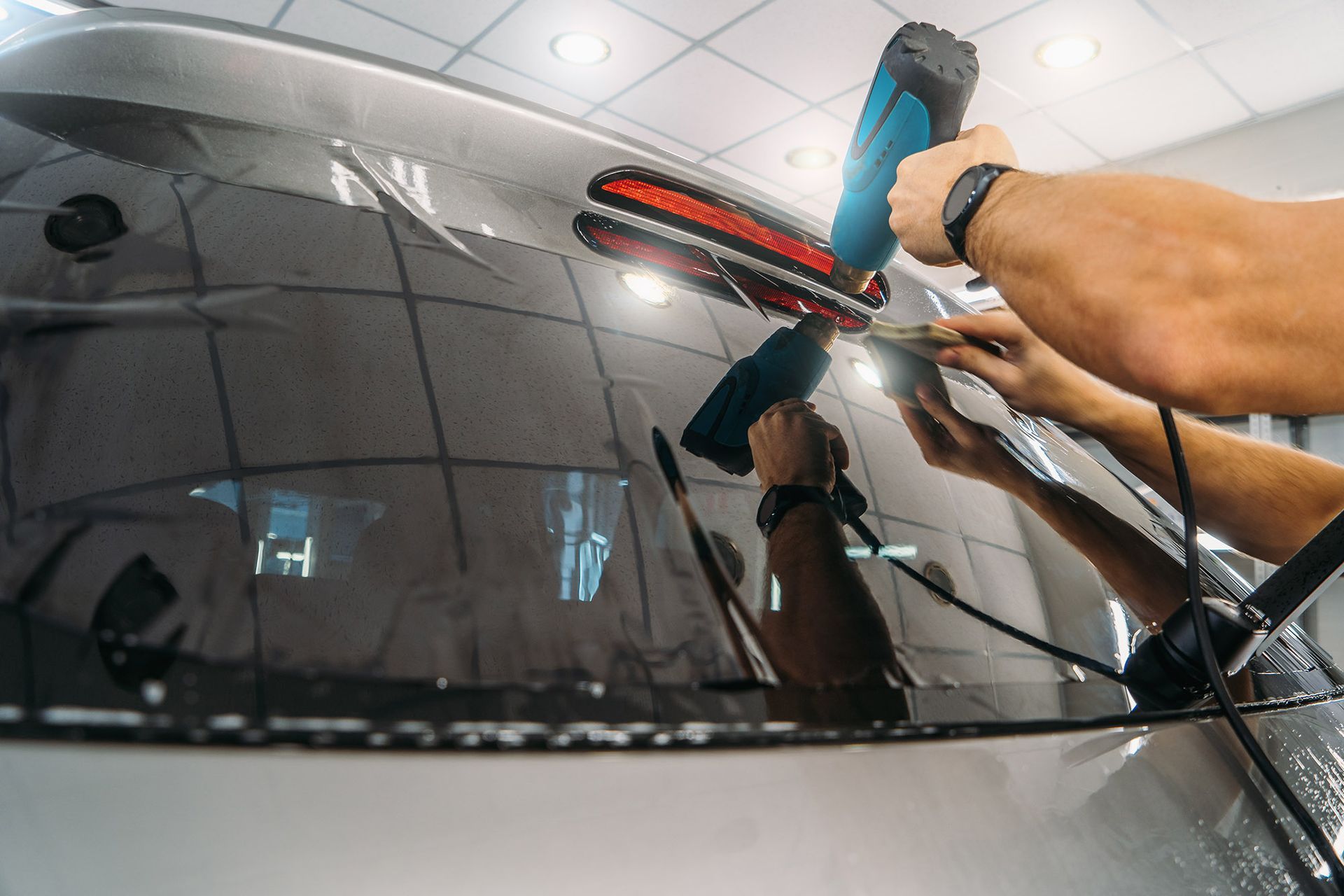 A technician uses a heat gun to apply window tinting film to a car's rear windshield inside a workshop.
