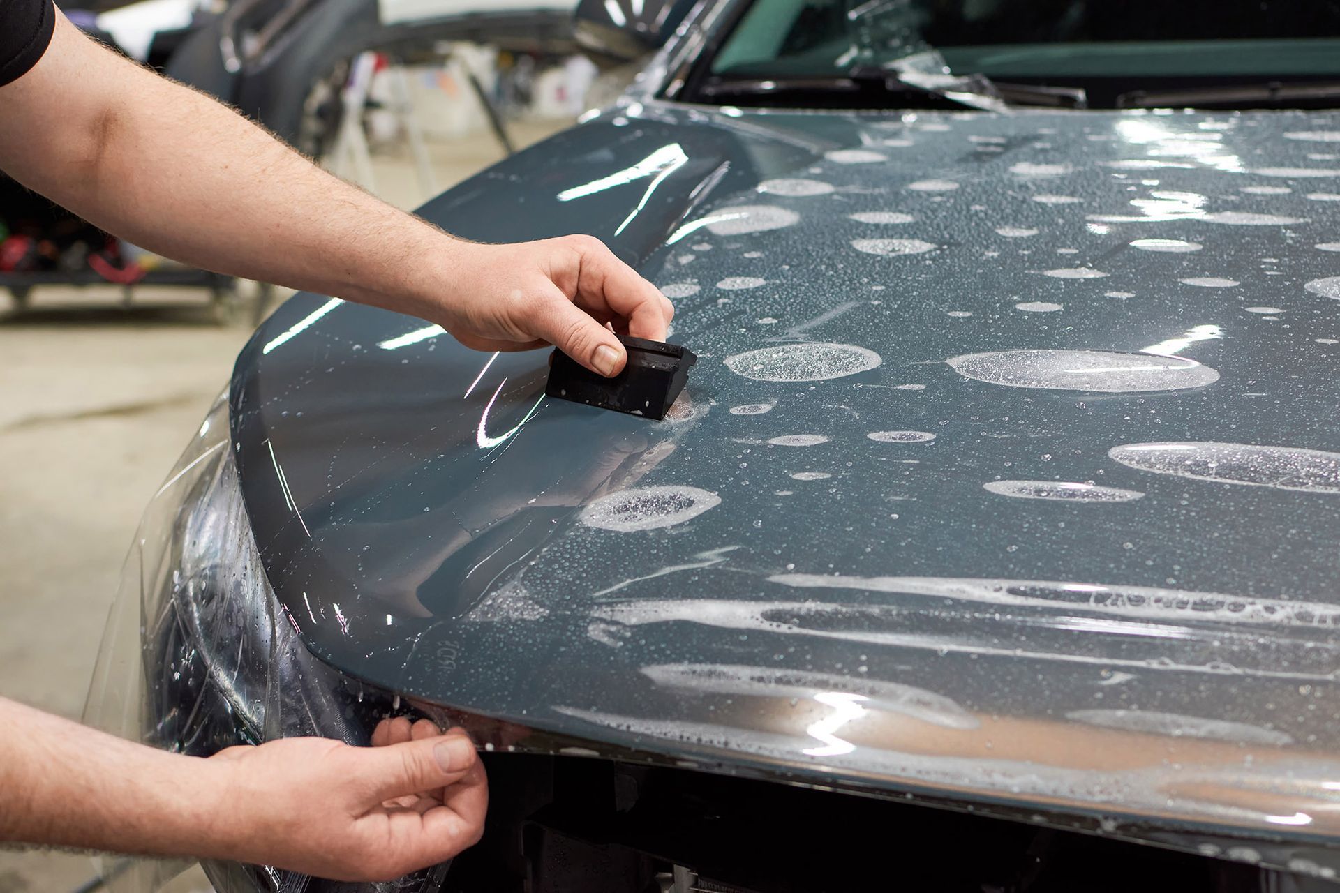 A person using a squeegee to apply a clear protective film to the gray hood of a car.