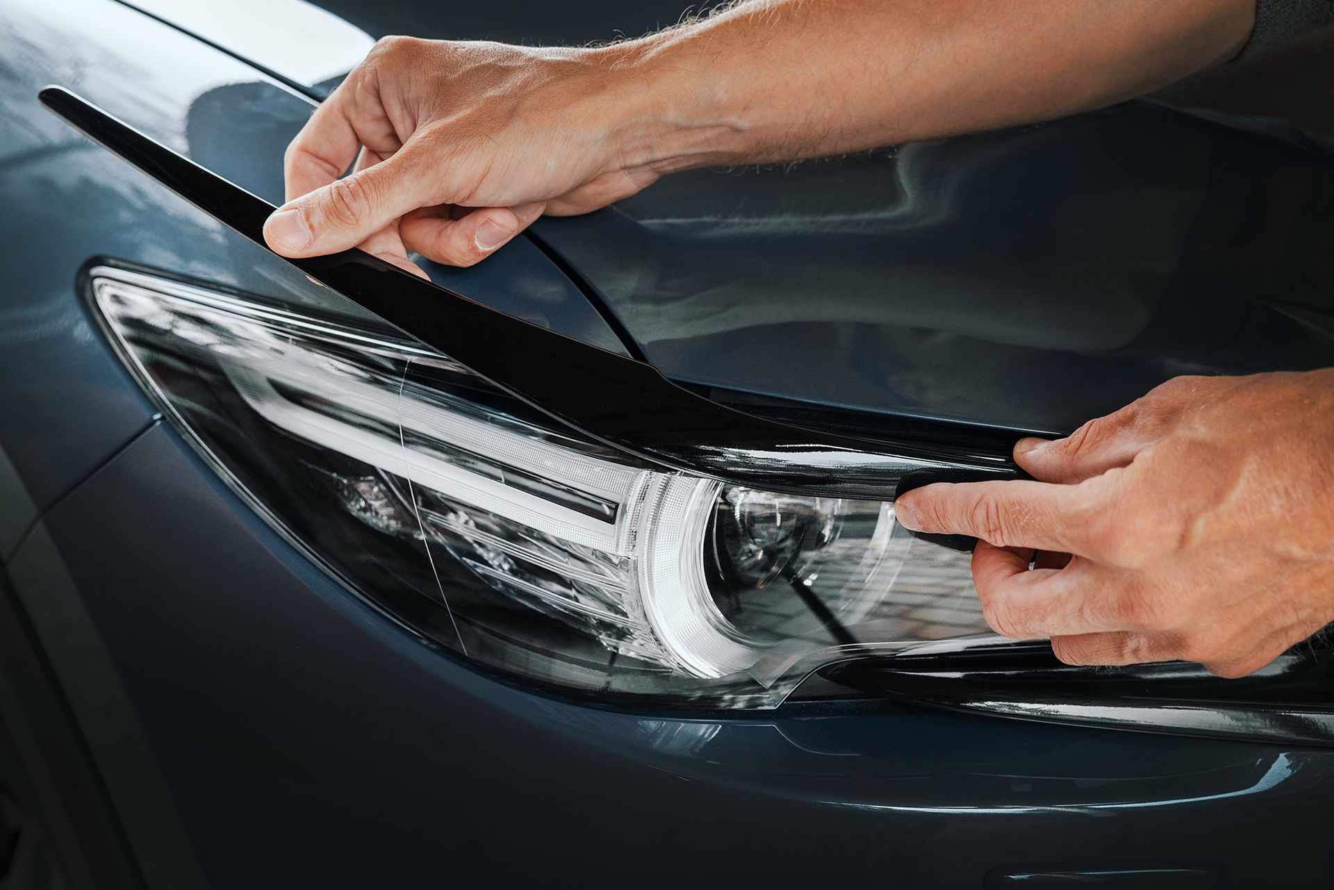 Hands applying a protective clear film onto the headlight of a dark-colored car.