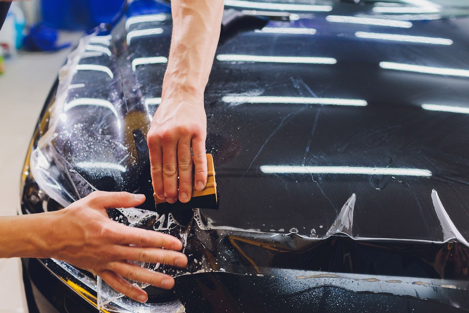 A person uses a squeegee to apply a clear protective film to the black hood of a car.