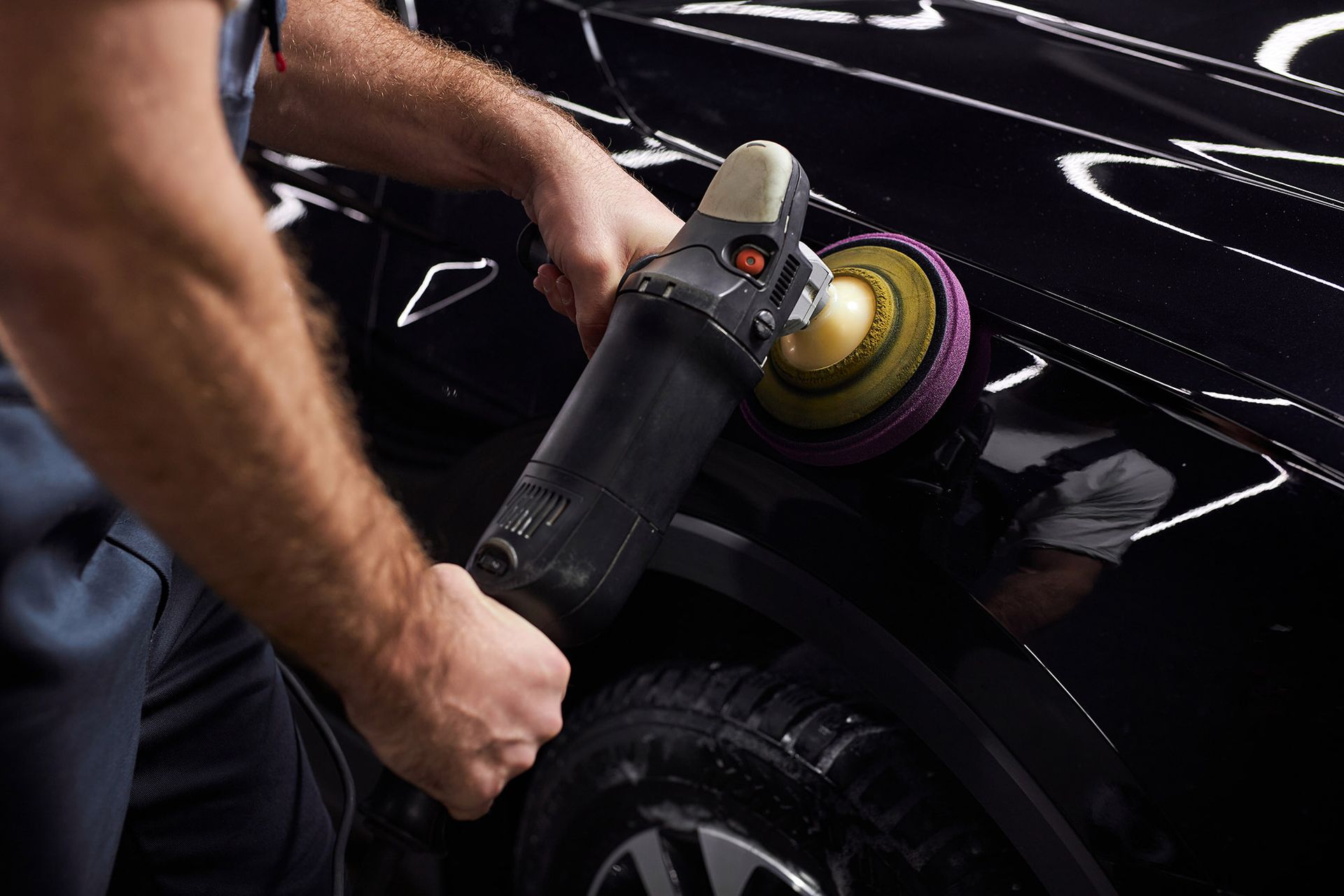 A person uses a rotary buffer tool to polish the black fender of a car in a workshop.