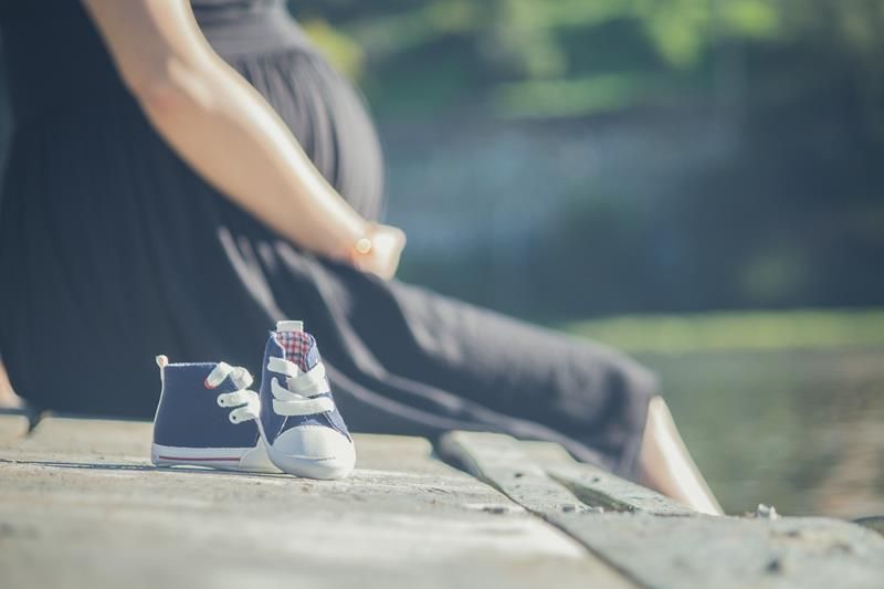 Pregnant person in black dress sits by water, baby sneakers in front.