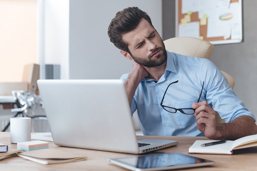 Man at desk holding glasses, rubbing neck, looking at laptop, indoors.