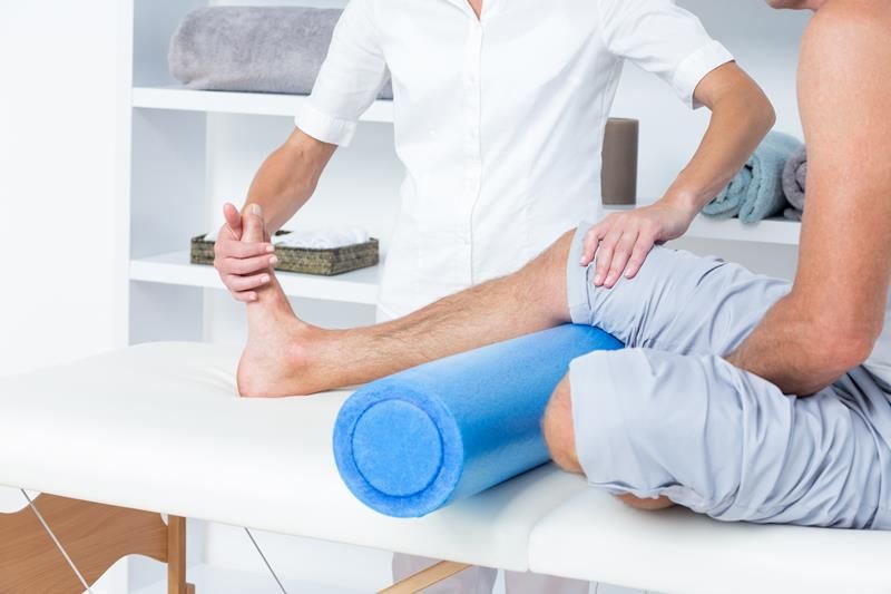 Therapist stretching a person’s leg on a treatment table, using a foam roller for support in a white room.