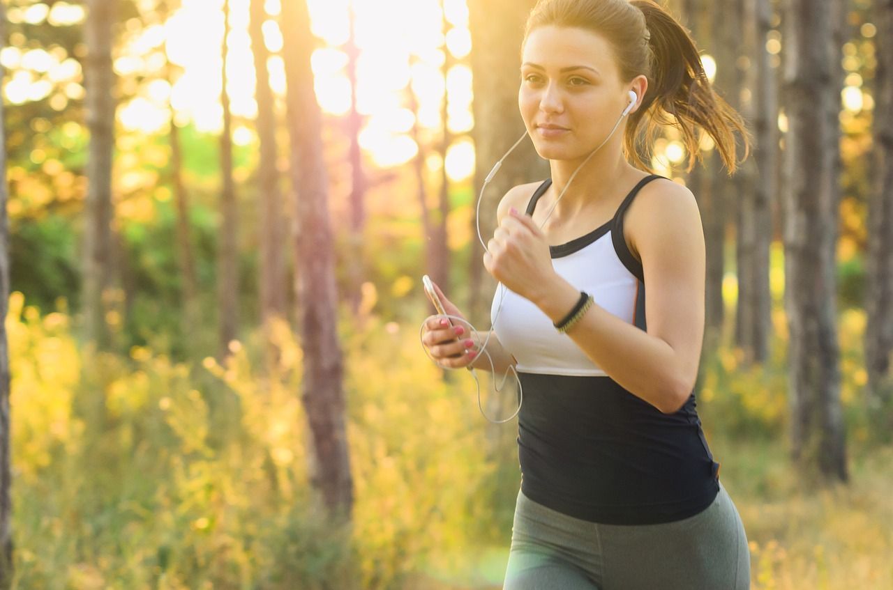 Woman running through a sunlit forest, wearing earbuds, holding phone.